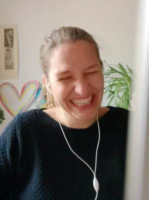 Julia Harms, Grafikdesignerin aus Nürnberg, mit dunkelblondem Haar, die einen dunklen Pullover und weiße Kopfhörer trägt, lacht breit mit geschlossenen Augen; im Hintergrund sind eine Pflanze und eine Herzzeichnung zu sehen, im Bildvordergrund der angeschnittene Bildschirm, der auf eine Telefonkonferenz hindeutet.