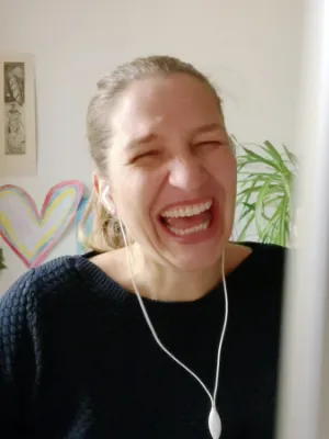 Als Grafikdesignerin Nürnberg bleibt Julia Harms im Austausch mit ihren Kund:innen - hier in einer Onlinekonferenz, erkennbar am angeschnittenen Bildschirm und den weißen in-ear-Kopfhörern. Die Frau mit dunkelblondem Pferdeschwanz und dunkelblauem Pulli lacht herzhaft.