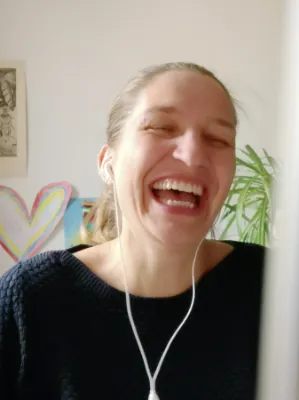 Als Grafikdesignerin Nürnberg bleibt Julia Harms im Austausch mit ihren Kund:innen und lacht bei einer Telefonkonferenz in den Bildschirm: im Hintergrund verschiedene Kunstwerke und Pflanzen.