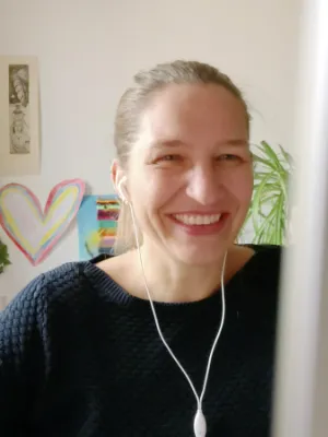 Grafikdesignerin Nürnberg: Julia Harms in der Telefonkonferenz mit Kund:innen. Sie trägt einen dunkelblauen Pulli und weiße in-ear-Kopfhörer mit Kabel. Im Vordergrund der angeschnittene Computerbildschirm, im Hintergrund Kunst und Kinder-Bildwerke.