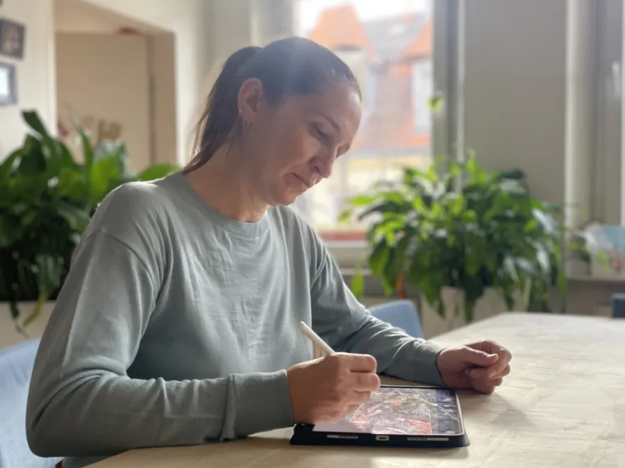 Grafikdesignerin Julia Harms, die ein hellblaues Sweatshirt trägt, zeichnet mit einem Stift auf einem Tablet, während sie an einem Tisch in einem hellen Raum mit Pflanzen im Hintergrund sitzt.