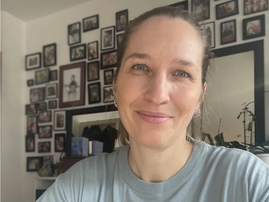 Eine Frau mit dunkelblondem Haar in einem Pferdeschwanz lächelt in die Kamera. Sie befindet sich in einem Raum, in dem im Hintergrund eine Wand mit gerahmten Fotos zu sehen ist.