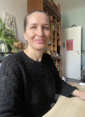 Eine Frau mit hellbraunem Haar und schwarzem Pullover sitzt lächelnd an einem Schreibtisch, mit Regalen und einer teilweise geöffneten Tür im Hintergrund.