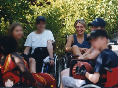 Eine Gruppe von Menschen, darunter einige in Rollstühlen, ist im Freien versammelt. Julia Harms, auch bekannt als julerennt, sitzt bei ihnen und lächelt breit strahlend in die Kamera.