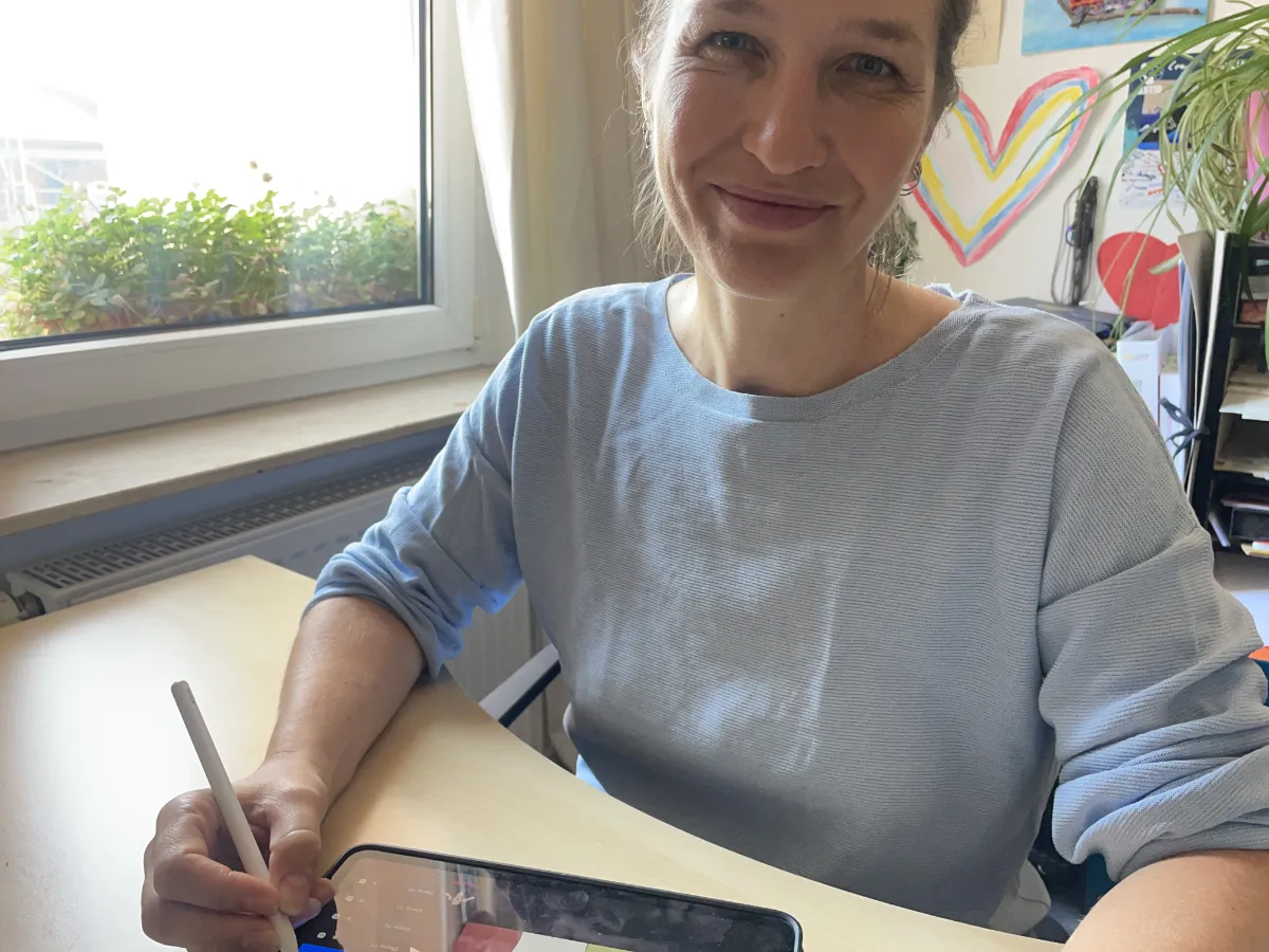 Eine Frau in einem hellblauen Hemd, Grafikdesignerin Julia Harms, sitzt an einem Schreibtisch am Fenster, lächelt und bedient mit einem Stylus ein Tablet. An der Wand im Hintergrund sind bunte Zeichnungen und eine Herzform zu sehen.