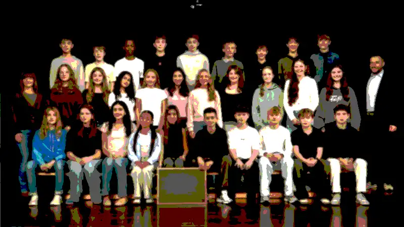 Eine Gruppe von Schülern und ein Erwachsener posieren für ein Klassenfoto in mehreren Reihen vor einem dunklen Hintergrund.